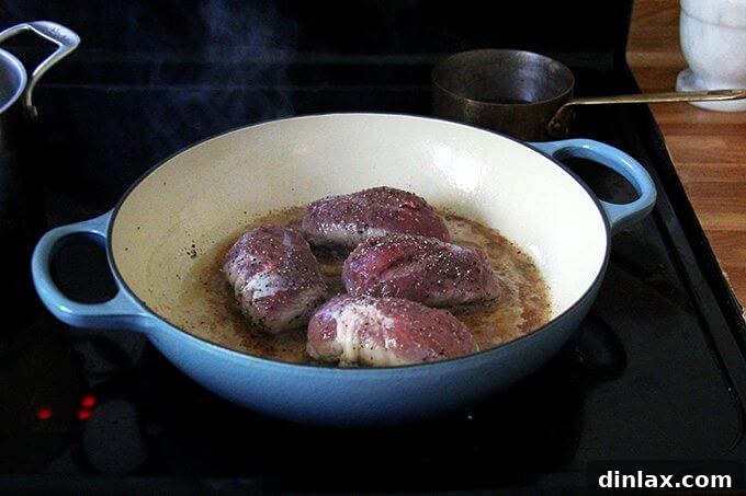 Four duck breasts cooking in a large Le Creuset braiser on the stovetop, fat rendering and skin browning.