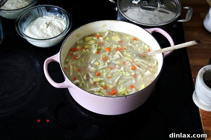 An overhead view of a stovetop, showing a large pot of cabbage soup with chopped carrots, potatoes, and other vegetables being added, while two bowls of rising bread dough rest nearby.