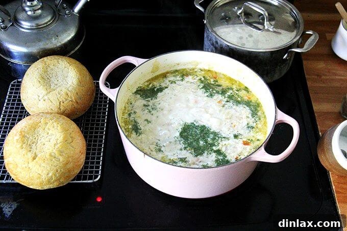 A large pot of vegetarian cabbage soup on the stovetop, showcasing the addition of heavy cream and finely chopped fresh dill, with two loaves of homemade bread resting nearby.