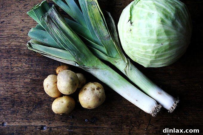 A wooden cutting board with a fresh head of cabbage, potatoes, and leeks, ready for chopping for homemade vegetable soup.