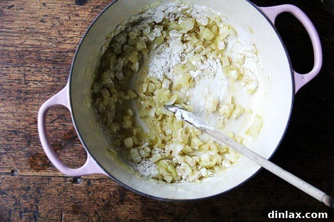 A large pot on the stovetop, containing sautéed onions now coated with flour, being stirred to form a roux for soup thickening.
