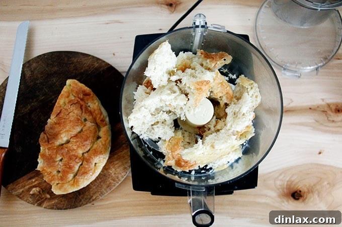 Roughly torn pieces of day-old focaccia bread placed inside the bowl of a food processor, ready to be processed into crumbs.