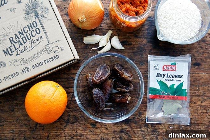 Instant Pot Harissa Rice with Sweet Dates and Zesty Orange 3 A selection of fresh ingredients laid out on a wooden board, including onions, garlic, a container of harissa paste, dried dates, a cup of basmati rice, and a whole orange.