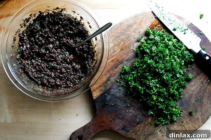 Zesty Olive Tapenade with Capers and Fresh Parsley 6 A bowl of black olive tapenade positioned next to a chopping board with freshly chopped parsley and a chef's knife.