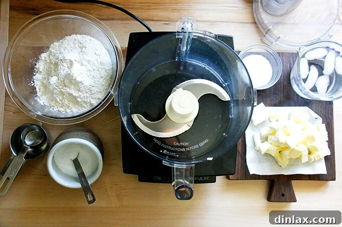 Rustic Rhubarb Frangipane Galette 5 A clean kitchen counter displaying a food processor with its blade, alongside bowls of all-purpose flour, sugar, salt, and cubes of cold butter – all ingredients for the foolproof pastry dough.