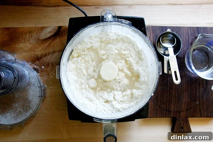 Rustic Rhubarb Frangipane Galette 9 Ice water being poured from a measuring cup into the food processor bowl through the feed tube, where dry ingredients and butter have been processed.