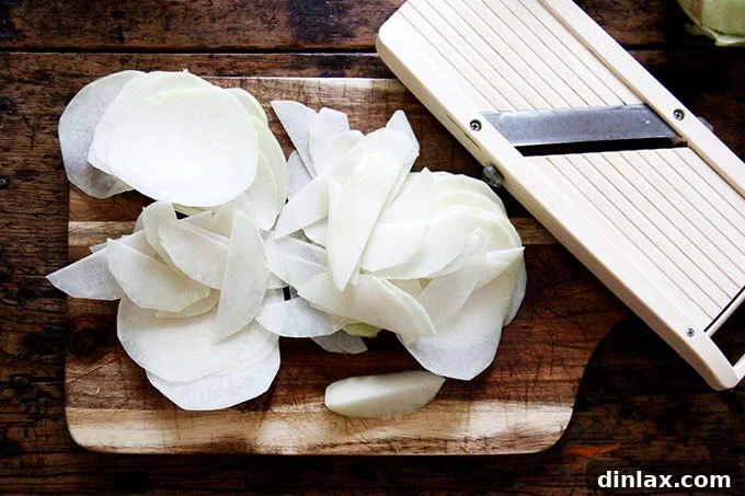 Thinly shaved kohlrabi on a wooden board next to a mandoline slicer, demonstrating the ideal slice thickness.