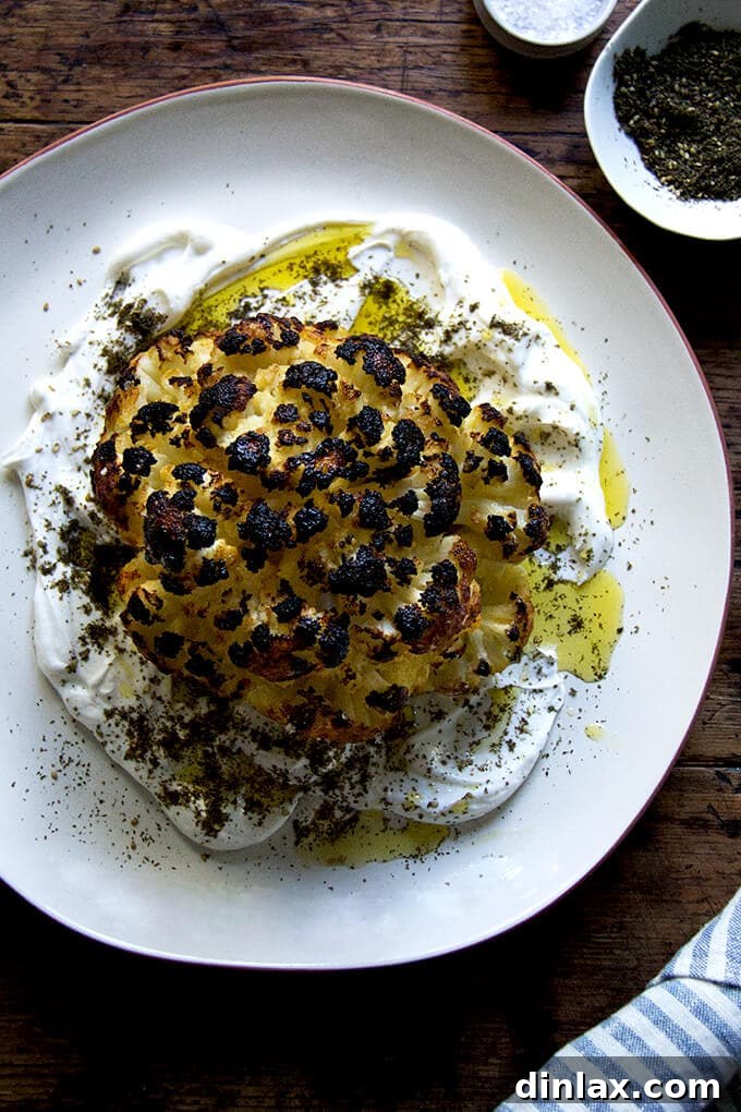 A beautifully roasted whole cauliflower head with golden-brown florets, drizzled with a creamy, vibrant lemony yogurt sauce and a sprinkle of za'atar, served on a rustic plate.
