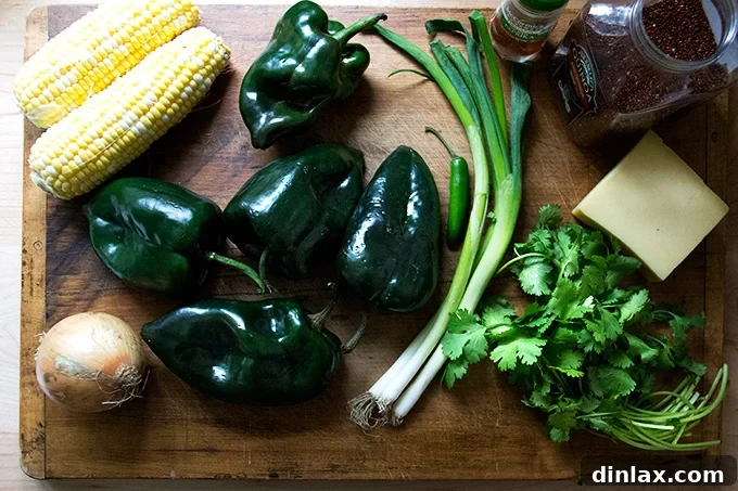 A cutting board displays fresh ingredients: vibrant corn kernels, whole poblano peppers, a halved onion, bright green scallions, fresh cilantro, a red jalapeño chili, uncooked quinoa, and blocks of Monterey Jack cheese.