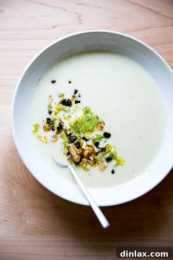 An overhead shot of a luxurious bowl of cream of celery soup generously topped with a vibrant walnut-currant salsa, garnished with fresh celery leaves.