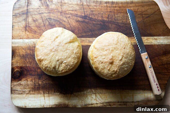 Two small, freshly baked loaves of bread on a wooden board next to a serrated knife, ready to be transformed into bread bowls for a rustic presentation.