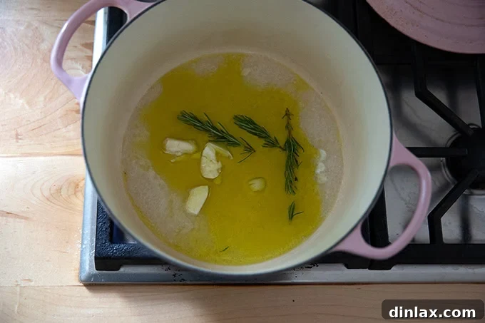 A large pot on the stovetop, containing a blend of olive oil, water, fresh rosemary sprigs, and smashed garlic cloves, ready for cooking.
