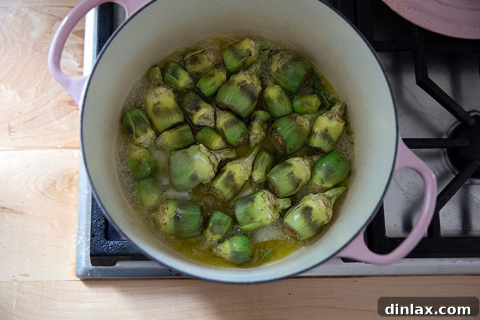 A large pot on the stovetop, now filled with baby artichokes gently cooking in the olive oil, water, rosemary, and garlic mixture.