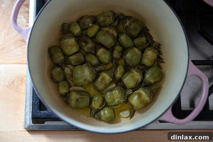 A large pot on the stovetop, with baby artichokes visibly softened and halfway through their covered cooking process.