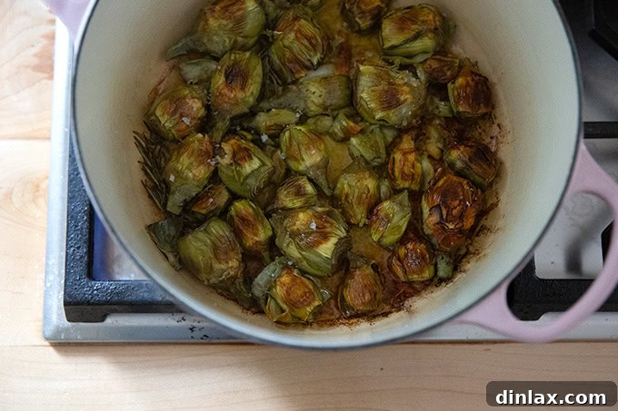 A large pot on the stovetop, revealing baby artichokes with one side perfectly crisped and golden brown, ready to be flipped.