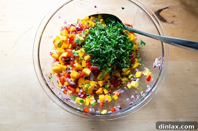 Freshly chopped cilantro being added to a large bowl of mango salsa, creating a vibrant contrast of green against the colorful fruit and vegetables.