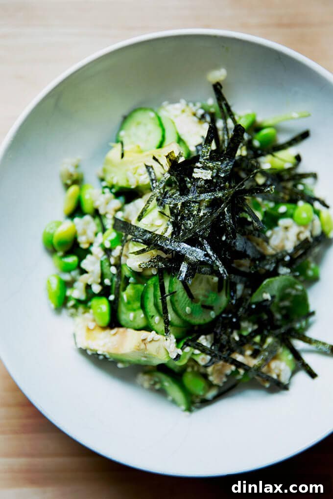 A vibrant bowl of sushi salad featuring nutty brown rice, creamy avocado, crisp edamame, and fresh cucumber, all drizzled with a rich sesame miso dressing and garnished with delicate nori strips and toasted sesame seeds.