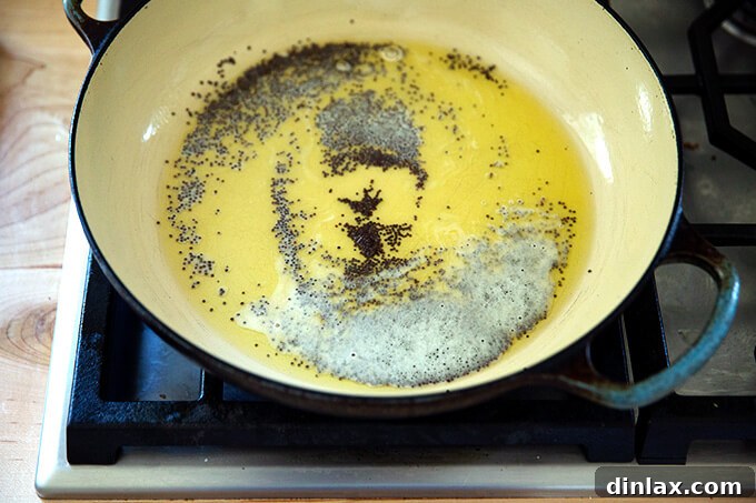 A braiser pan on the stovetop, heating fragrant ghee with an initial sizzle of black mustard seeds, beginning the tempering process.