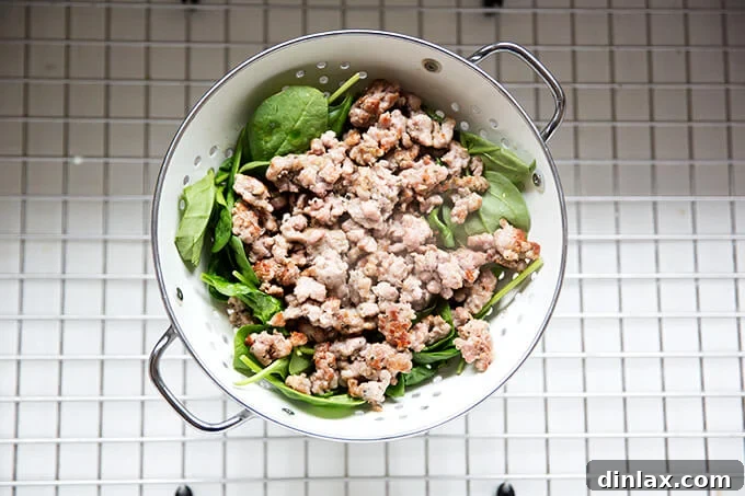 Sausage and fresh spinach draining and gently wilting together in a colander in the sink.