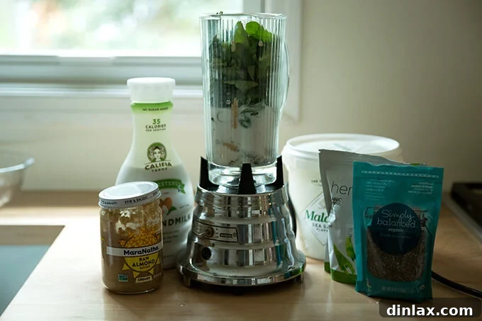 All of the ingredients for the green smoothie on the counter ready to be blended in the blender. Fresh spinach, frozen banana slices, almond milk, almond butter, dates, and ice.