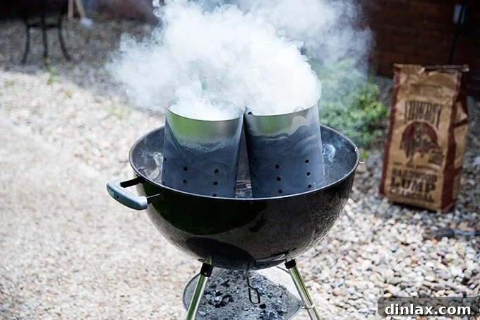 A Weber grill being prepared for grilling, with two smoking chimney starters filled with hot coals, demonstrating proper charcoal lighting technique.