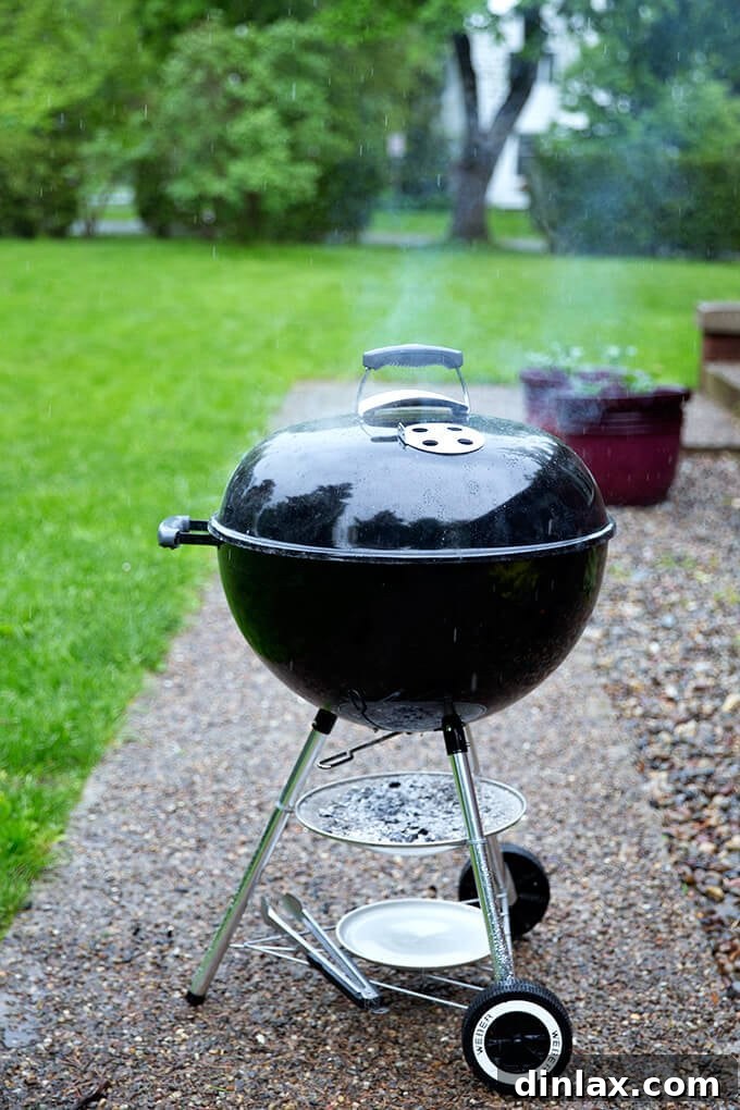 A dedicated griller tending to a Weber grill with hot coals, undeterred by light rain, proving that grilling can be an all-weather activity.
