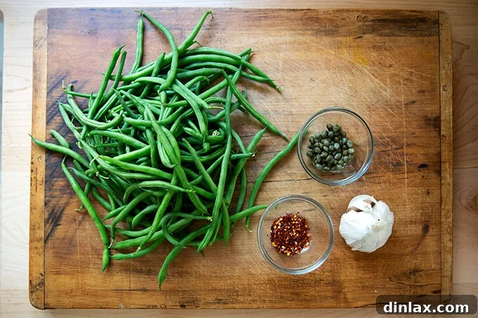 Various ingredients for the spicy, blistered green beans recipe including fresh green beans, garlic, capers, olive oil, and chili flakes.