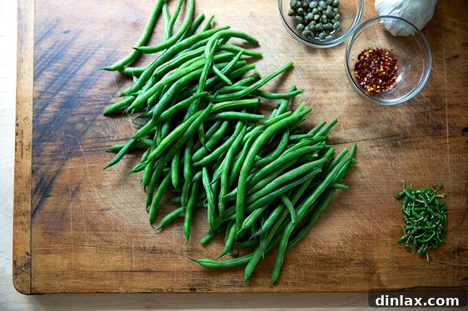 A cutting board showcasing prepped ingredients: stemmed green beans, a pile of capers, whole garlic cloves, and a small mound of chili flakes.