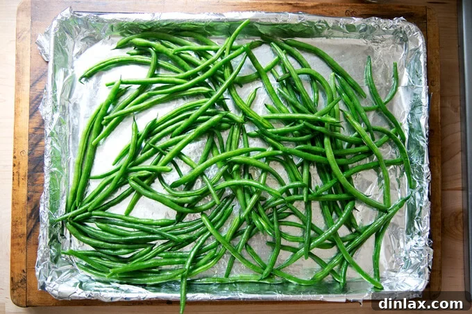A foil-lined sheet pan covered evenly with fresh green beans, prepped for broiling.