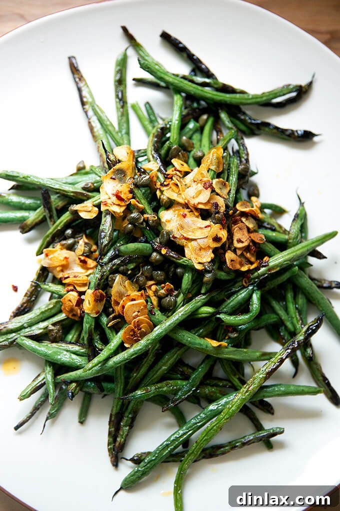 A close-up shot of a plate of spicy, blistered green beans with garlic and caper oil, highlighting the vibrant colors and charred textures.