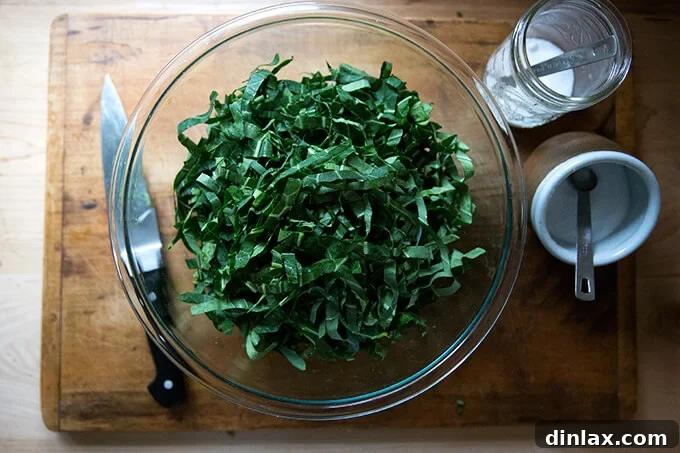 A bowl of thinly sliced raw collard greens, positioned next to small bowls containing kosher salt and granulated sugar, and a knife.