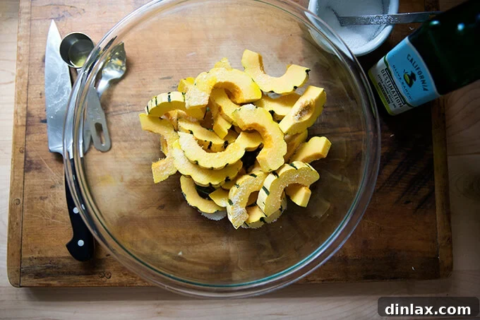 Raw delicata squash slices, tossed with olive oil and a sprinkle of salt, in a large mixing bowl, ready for roasting.
