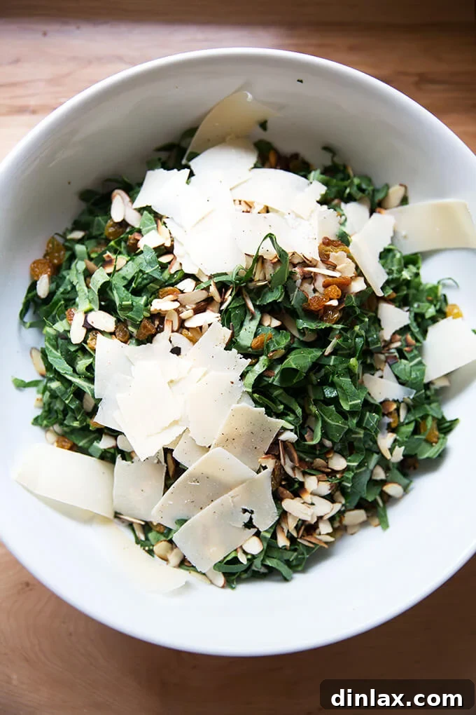 A large bowl containing thinly shaved collard greens, mixed with toasted almonds, golden raisins, and generous shavings of Parmesan cheese, awaiting the dressing.