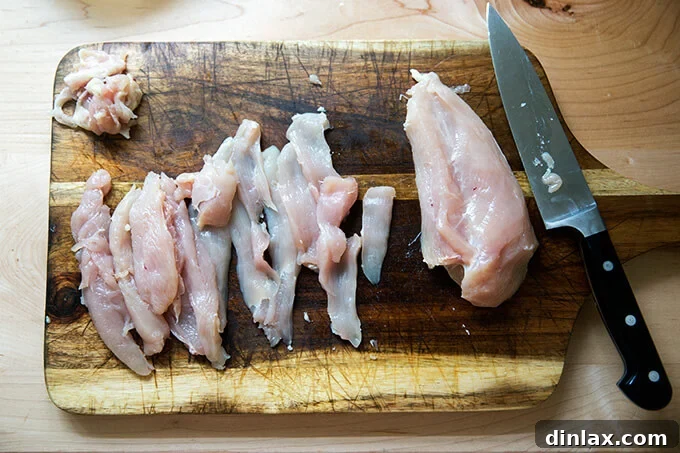 A clean cutting board displays several boneless, skinless chicken breasts, expertly thinly sliced into long, even strips, ready for marinating and skewering.