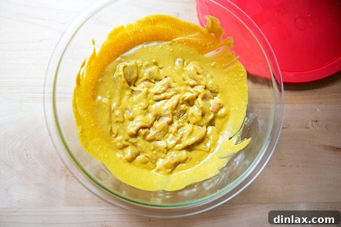 A clear glass bowl contains thinly sliced chicken breast strips generously coated in the rich, golden easy Thai chicken satay marinade, ready for refrigeration.