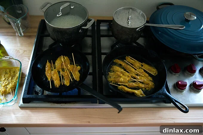 Two cast iron skillets are actively cooking easy Thai chicken satay skewers on a stovetop, with the chicken developing a beautiful golden crust.