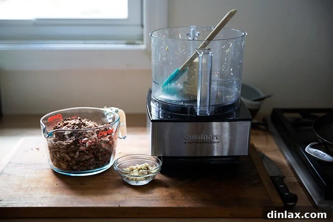 An empty food processor sits beside a liquid measuring cup filled with finely chopped mushrooms and minced ginger, prepped for cooking.