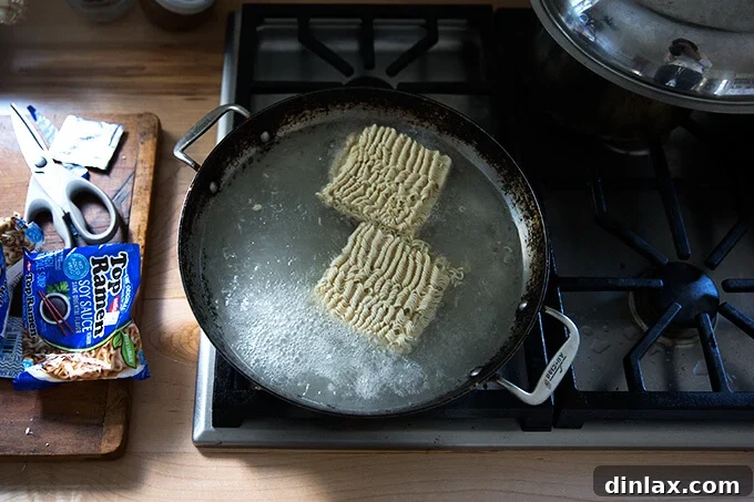 A large sauté pan on the stovetop, filled with boiling water and ramen noodles, beginning to cook.