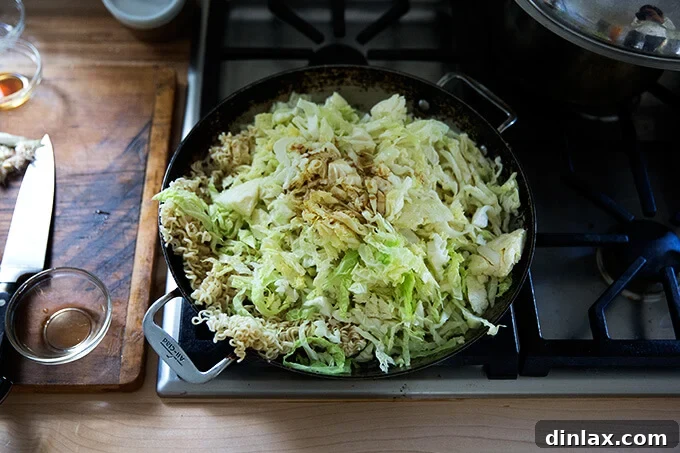 A large sauté pan filled with sautéed mushrooms, fragrant ginger, wilted cabbage, and par-cooked ramen noodles, ready to be tossed.