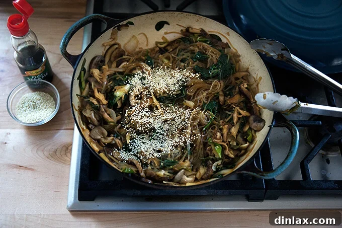 Japchae: Vegetarian Korean Glass Noodles 15 Freshly cooked japchae in a braiser on the stovetop, being garnished with sesame seeds and a drizzle of sesame oil.