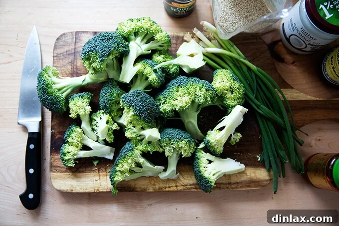 A cutting board displays fresh broccoli florets and spears alongside essential ingredients for the sizzle oil: sesame oil, coconut oil, soy sauce, Sambal Oelek, and green onions, illustrating the preparation for spicy broiled broccoli.