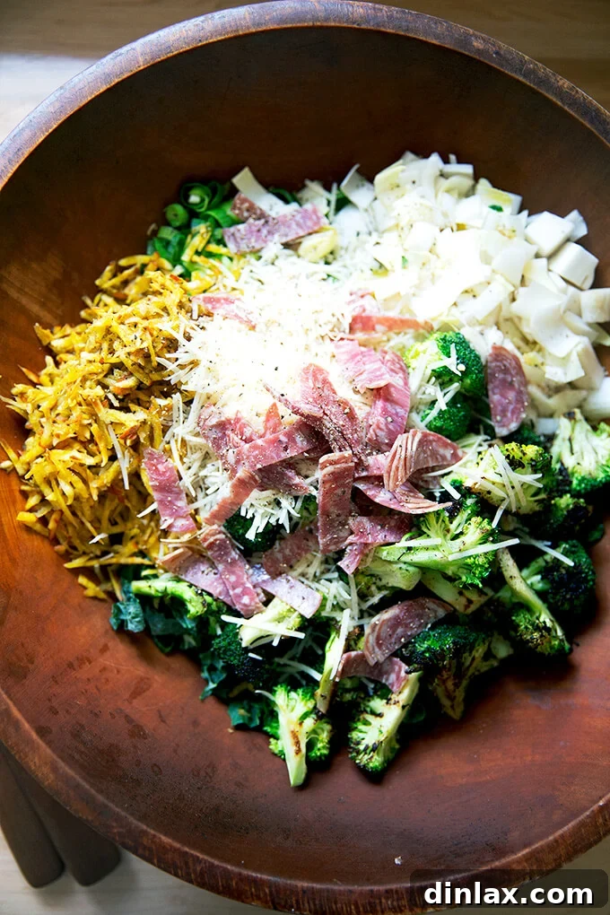 A large salad bowl filled with all the chopped salad ingredients, including vegetables, salami, and cheese, before dressing.