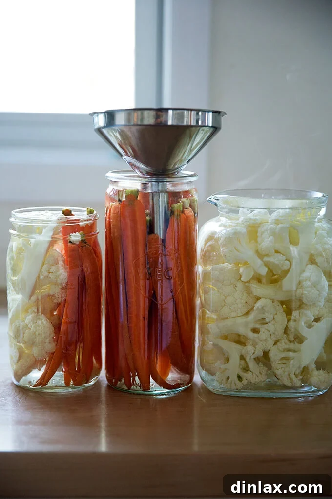 Glass jars of crudités, one with a funnel, being filled with warm pickling liquid.