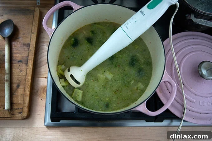Golden Cheddar Broccoli Bisque 9 An immersion blender is actively puréeing the broccoli-cheddar soup directly in the Dutch oven, creating a smooth and creamy texture.