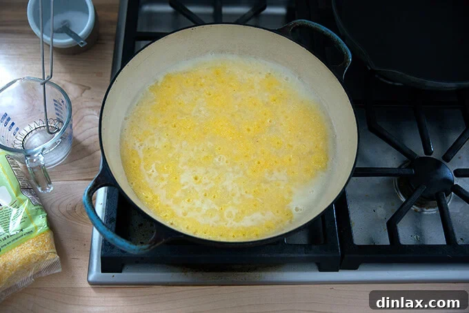 A large skillet on the stovetop filled with polenta simmering gently, with small bubbles breaking the surface.