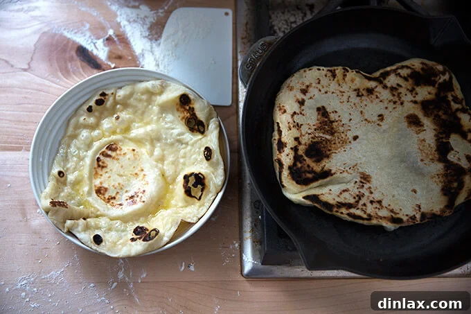 A freshly cooked naan being removed from a hot skillet, next to a stack of already cooked naan.