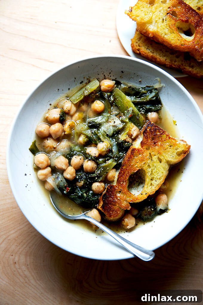 A close-up of a bowl of chickpea and escarole soup with a slice of toasted bread, generously sprinkled with fresh black pepper.
