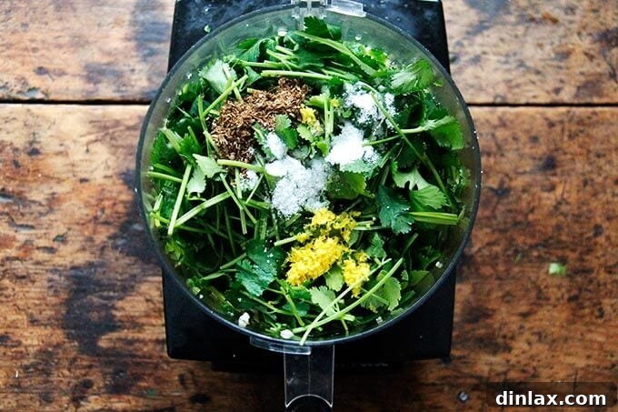 A food processor bowl filled with fresh cilantro, parsley, ground spices, lemon zest, garlic, and chilies, before final processing.