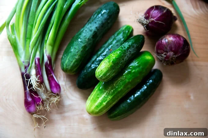 Fresh scallions, cucumbers, and red onions arranged on a wooden cutting board, ready for preparation.