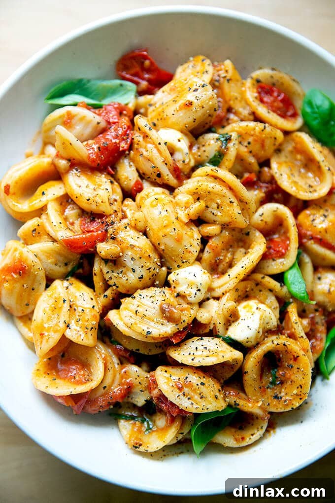 A vibrant bowl of orecchiette pasta generously tossed with bright red cherry tomato sauce, golden pine nuts, fresh green basil leaves, and creamy white mozzarella pearls, garnished with shaved Parmesan.